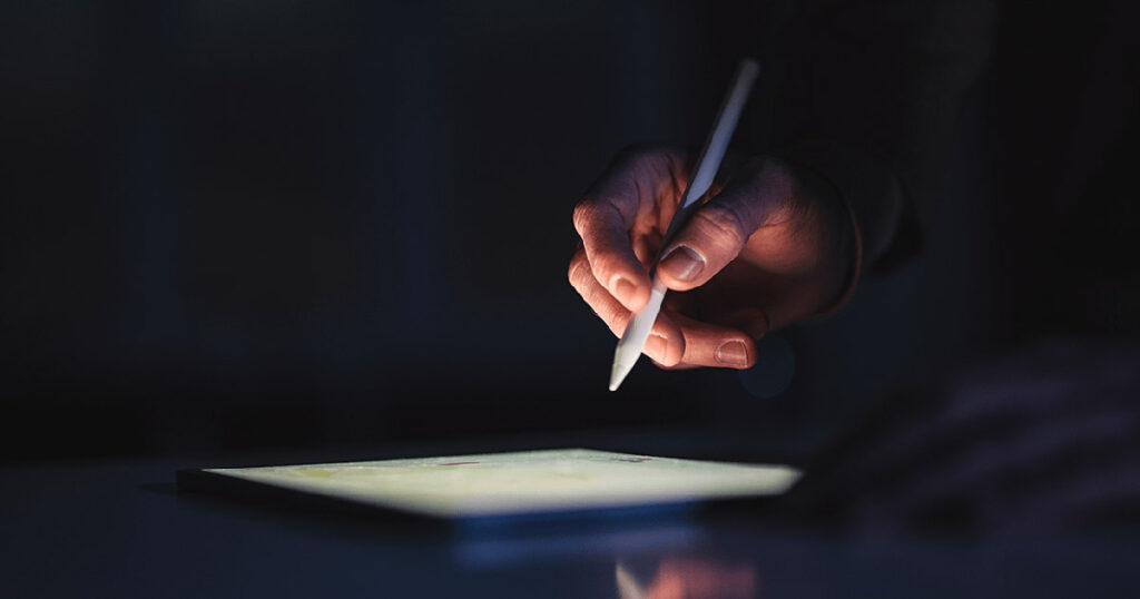 A hand holding a pen on a tablet in the dark.