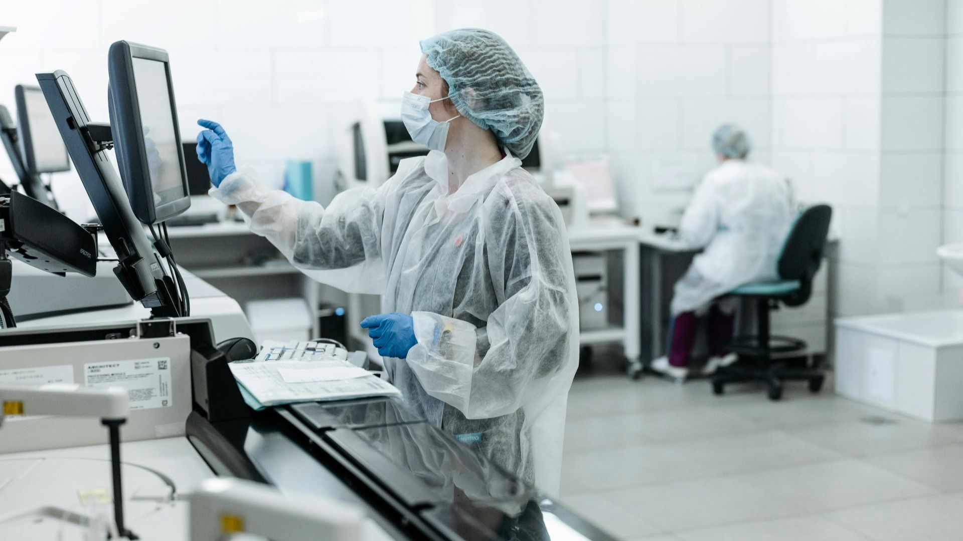 Two lab technicians wearing protective gear work in a laboratory within the pharmaceutical industry; one is using a touchscreen monitor, while the other sits at a desk in the background.