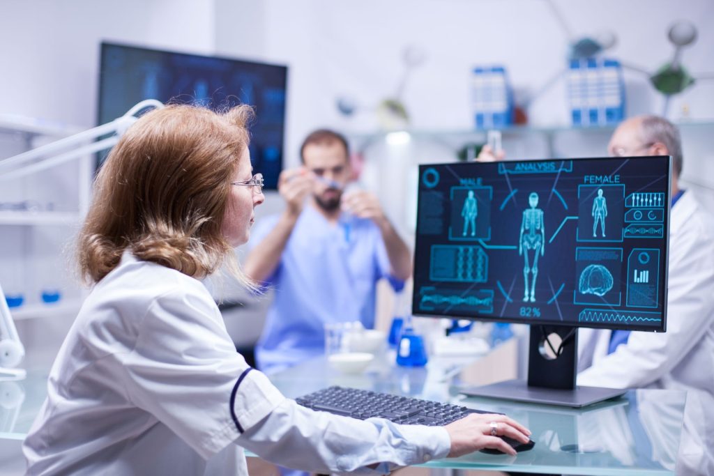 A scientist works on a computer displaying medical data. In the background, two other scientists conduct experiments in a lab. The environment is bright and clinical.