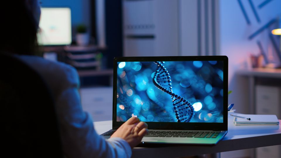 A person sits at a desk looking at a laptop displaying an image of a DNA double helix, indicative of multi-omics data analysis. The background shows office equipment.