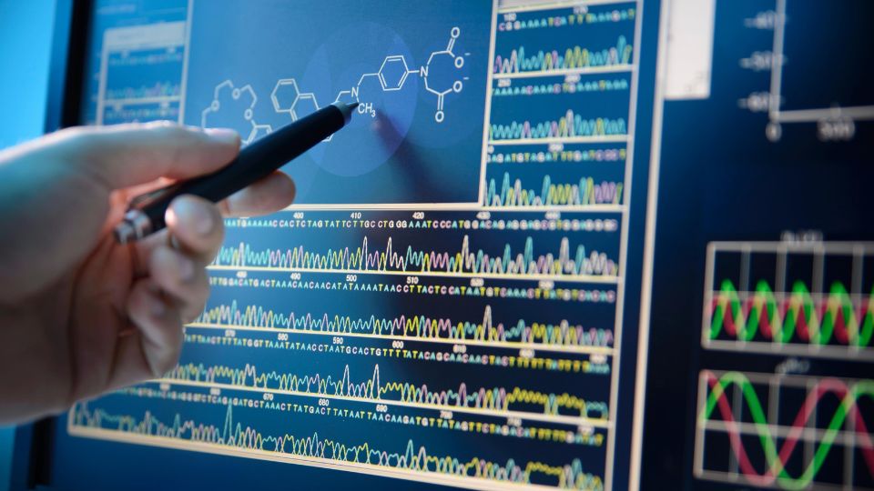 A hand holding a pen points at molecular structure and multi-omics data displayed on a computer screen.