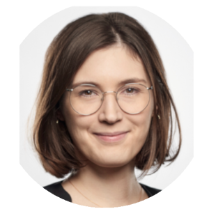 Portrait of a person with shoulder-length brown hair, glasses, and a slight smile, wearing a black top against a plain background.