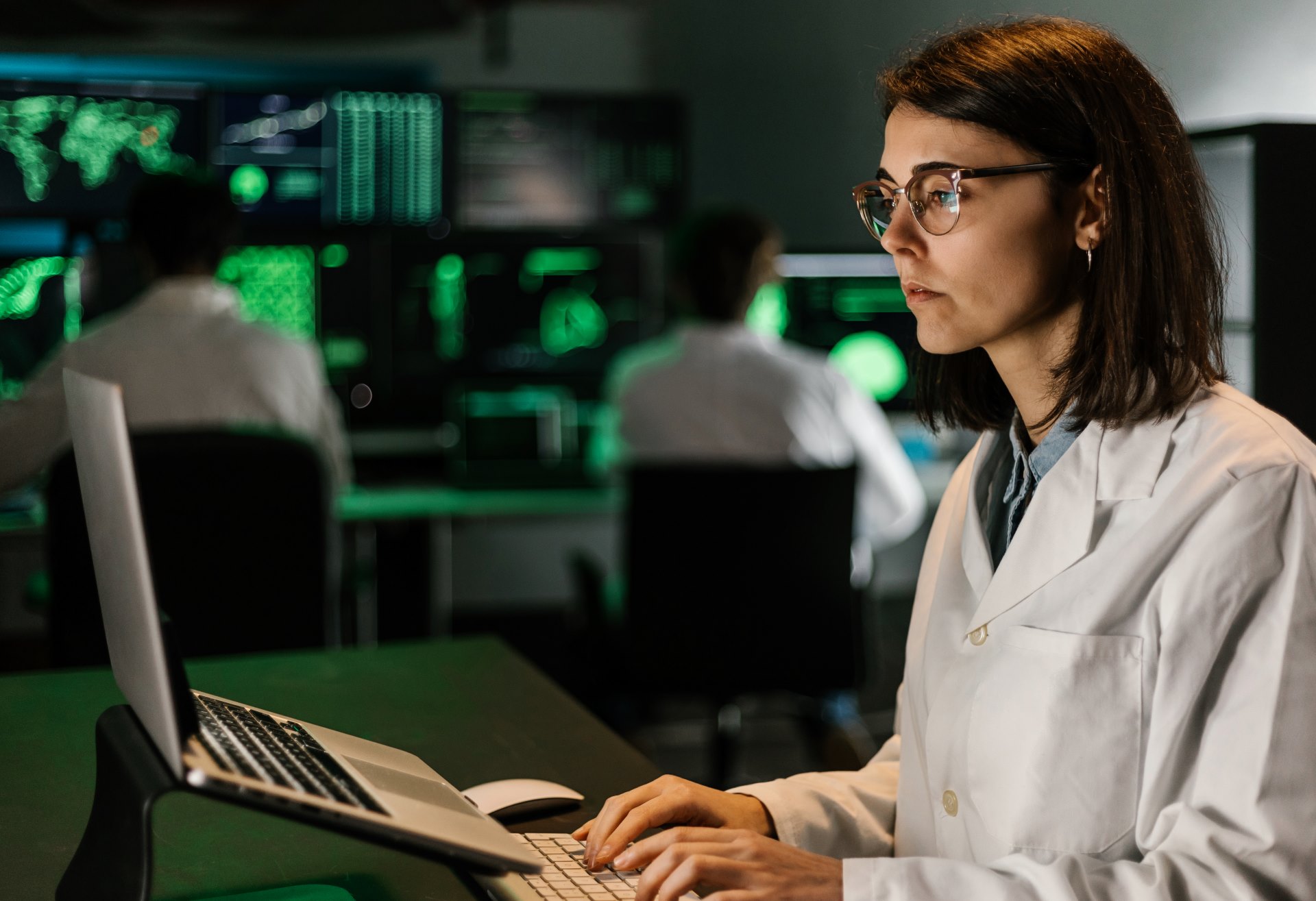 A woman in a lab coat and glasses works on a laptop in a dimly lit room with multiple screens displaying data in the background, as part of the EU Horizon Research initiative. Other individuals also work at computers behind her, contributing to the collaborative effort.