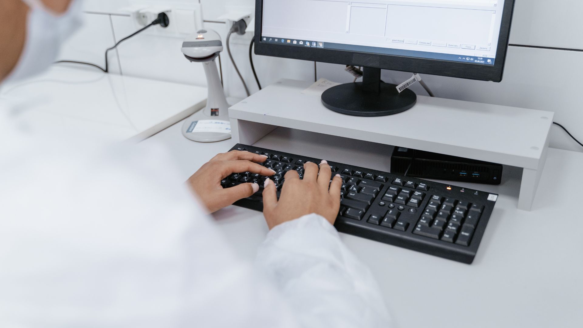 A person wearing a white lab coat works on a desktop computer keyboard in a laboratory setting with various equipment in the background.