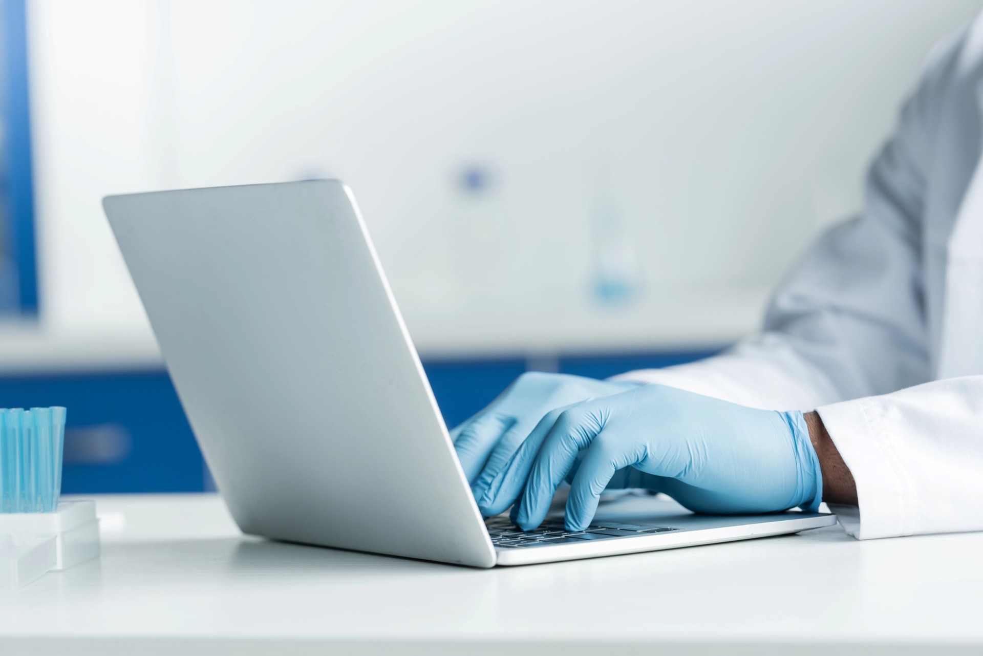 Person wearing blue gloves and a lab coat typing on a laptop in a laboratory setting.
