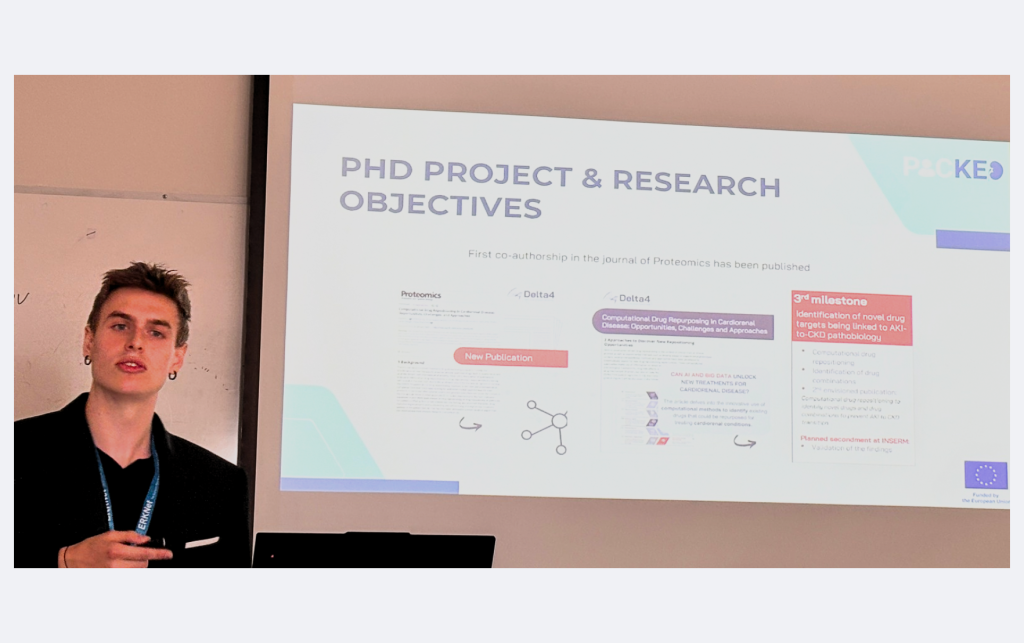 A man stands near a projection screen displaying a picked slide titled "PhD Project & Research Objectives," featuring text, diagrams, and highlighted sections.
