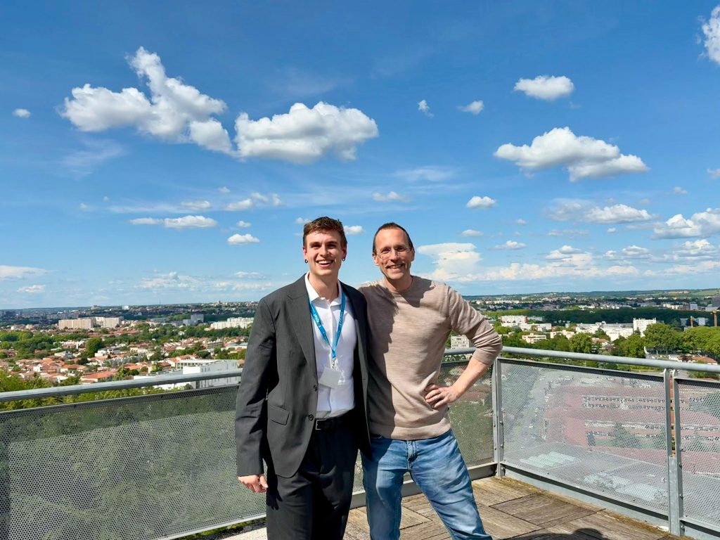 Two men stand on a balcony overlooking a cityscape and blue sky with scattered clouds. One picked a suit for the day, while the other chose a casual sweater and jeans.