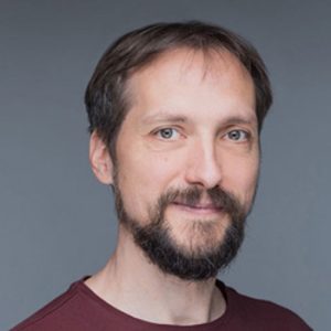 A member of Delta4's team with short brown hair and a beard, wearing a maroon shirt, smiles slightly in front of a plain gray background.
