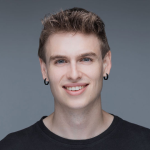 A young man with short, light brown hair, wearing a black shirt and black hoop earrings, smiles at the camera against a plain grey background.