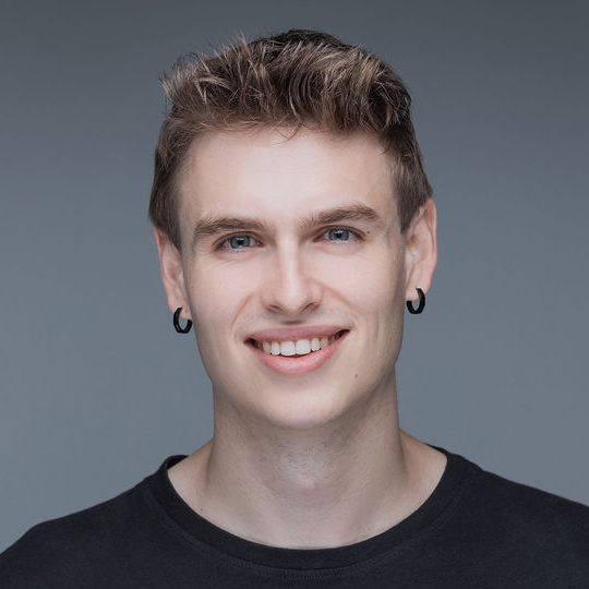 A young man with short, light brown hair, wearing a black shirt and black hoop earrings, smiles at the camera against a plain grey background.