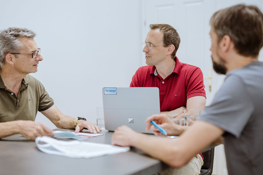 Three men sit at a table having a discussion; one has a laptop open, and documents and pens are on the table.