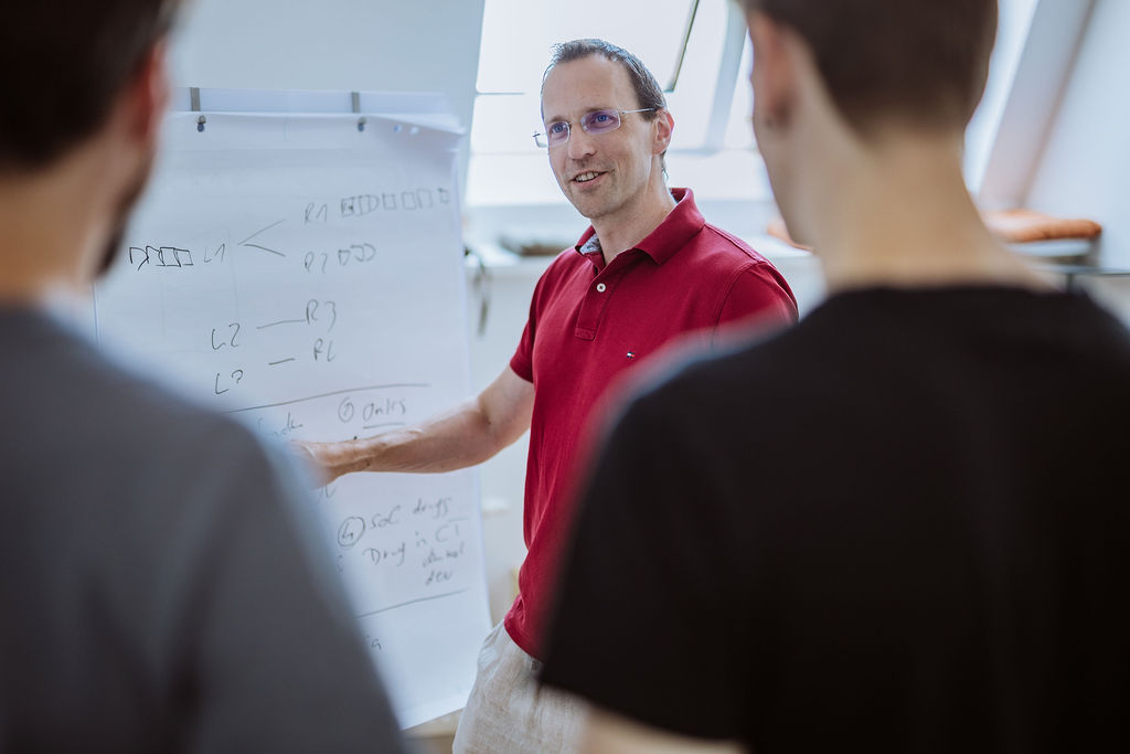 A man in a red polo shirt explains a diagram on a flip chart to two people in a well-lit room.