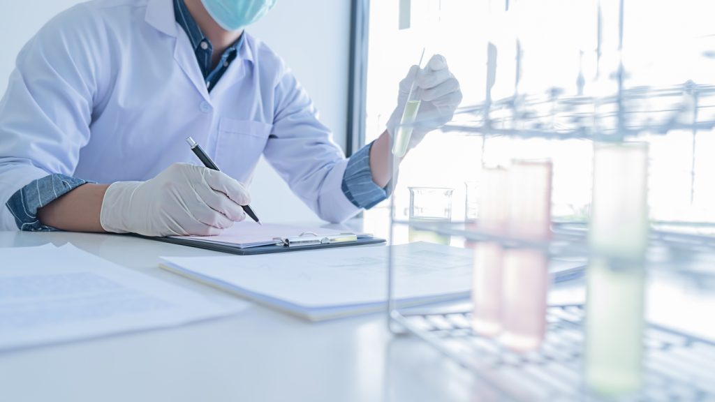 A scientist wearing gloves and a mask writes notes on a clipboard while handling a test tube beside a rack of test tubes in a laboratory setting.
