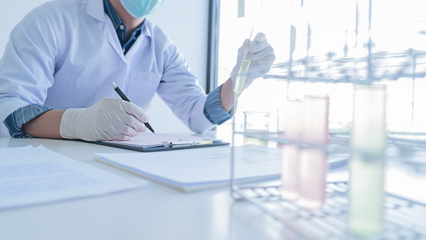 A scientist wearing gloves and a mask writes notes on a clipboard while handling a test tube beside a rack of test tubes in a laboratory setting.