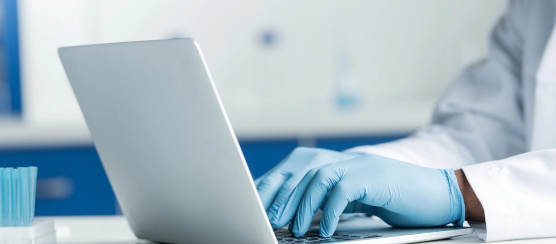 Person wearing blue gloves and a lab coat typing on a laptop in a laboratory setting.
