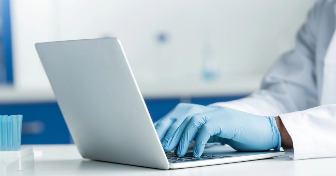 Person wearing blue gloves and a lab coat typing on a laptop in a laboratory setting.