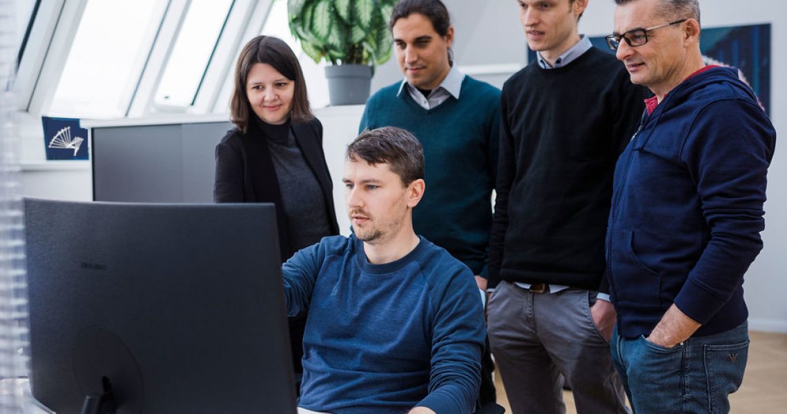 Five people gather around a desk, focusing on a computer screen. One person is seated and using the mouse, while the others stand behind observing. Indoor setting with natural light from large windows.
