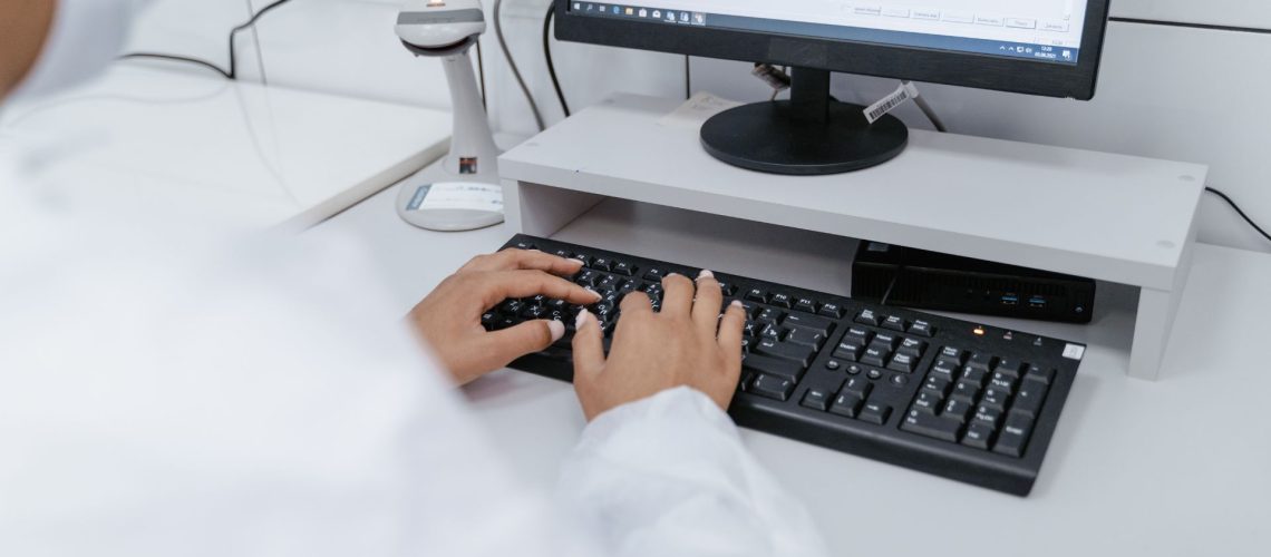 A person wearing a white lab coat works on a desktop computer keyboard in a laboratory setting with various equipment in the background.