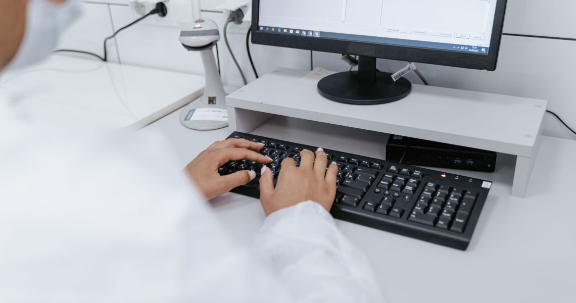 A person wearing a white lab coat works on a desktop computer keyboard in a laboratory setting with various equipment in the background.