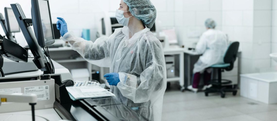 Two lab technicians wearing protective gear work in a laboratory within the pharmaceutical industry; one is using a touchscreen monitor, while the other sits at a desk in the background.