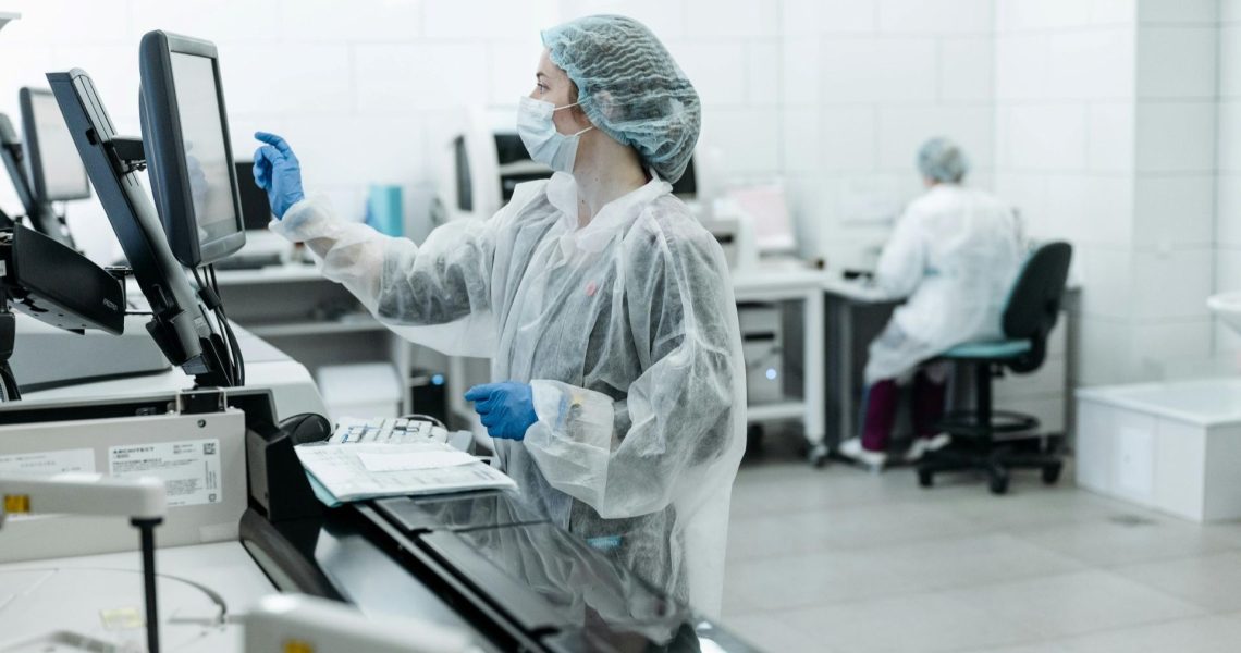 Two lab technicians wearing protective gear work in a laboratory within the pharmaceutical industry; one is using a touchscreen monitor, while the other sits at a desk in the background.