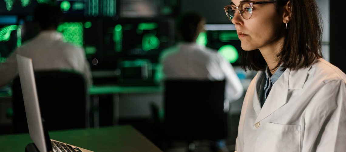 A woman in a lab coat and glasses works on a laptop in a dimly lit room with multiple screens displaying data in the background, as part of the EU Horizon Research initiative. Other individuals also work at computers behind her, contributing to the collaborative effort.