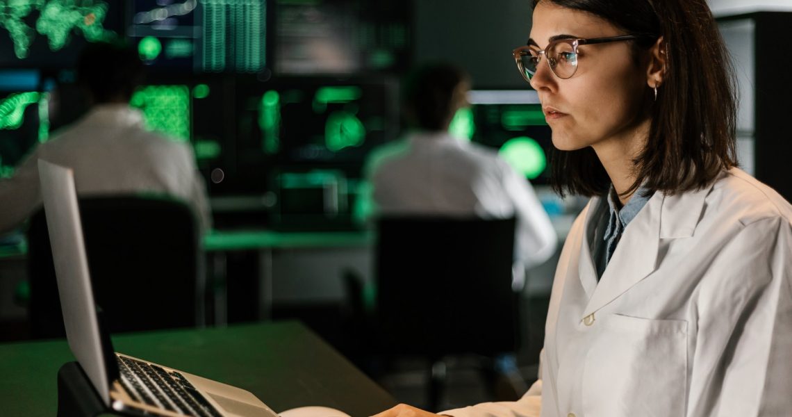 A woman in a lab coat and glasses works on a laptop in a dimly lit room with multiple screens displaying data in the background, as part of the EU Horizon Research initiative. Other individuals also work at computers behind her, contributing to the collaborative effort.