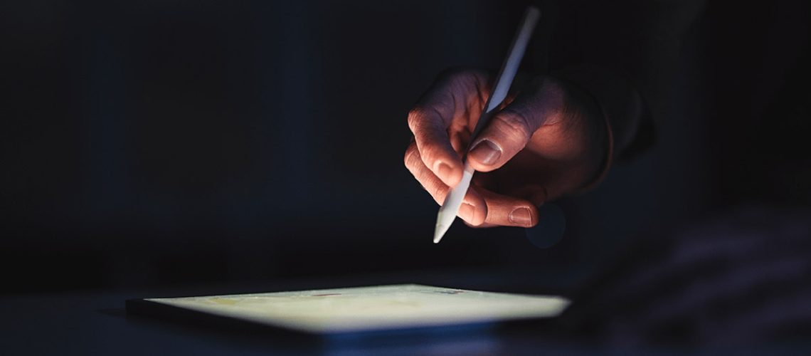 A hand holding a pen on a tablet in the dark.