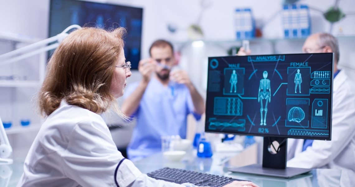 A scientist works on a computer displaying medical data. In the background, two other scientists conduct experiments in a lab. The environment is bright and clinical.